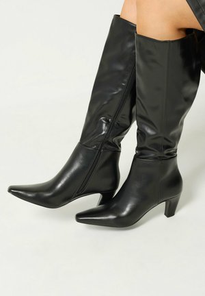 Bottes en cuir noires montantes jusqu'aux genoux avec des bouts pointus et des talons de hauteur moyenne, portées sur des jambes sur fond blanc.
