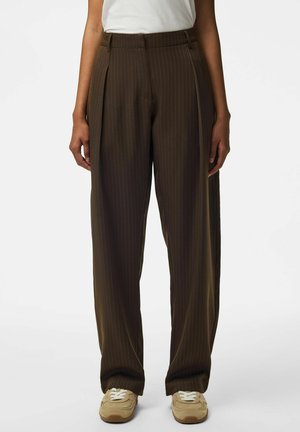 Personne portant un pantalon large marron à fines rayures, associé à des baskets beiges et un haut blanc sans manches, debout devant un fond uni.
