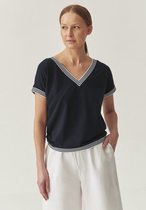 T-shirt marine avec un col en V et une bordure rayée blanche au niveau de l'encolure et des manches. Porté avec un pantalon ample de couleur claire.