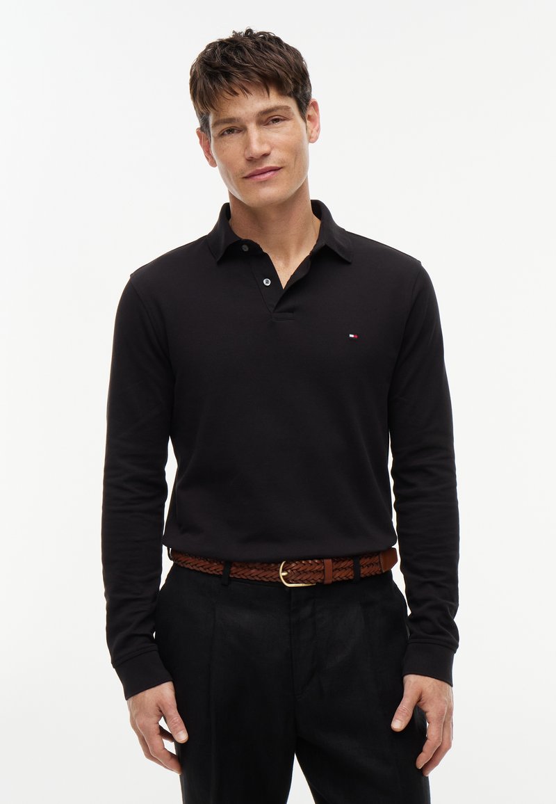 Hombre con pelo corto y castaño que lleva una camisa polo negra de manga larga, pantalones negros y un cinturón marrón tejido sobre un fondo blanco.