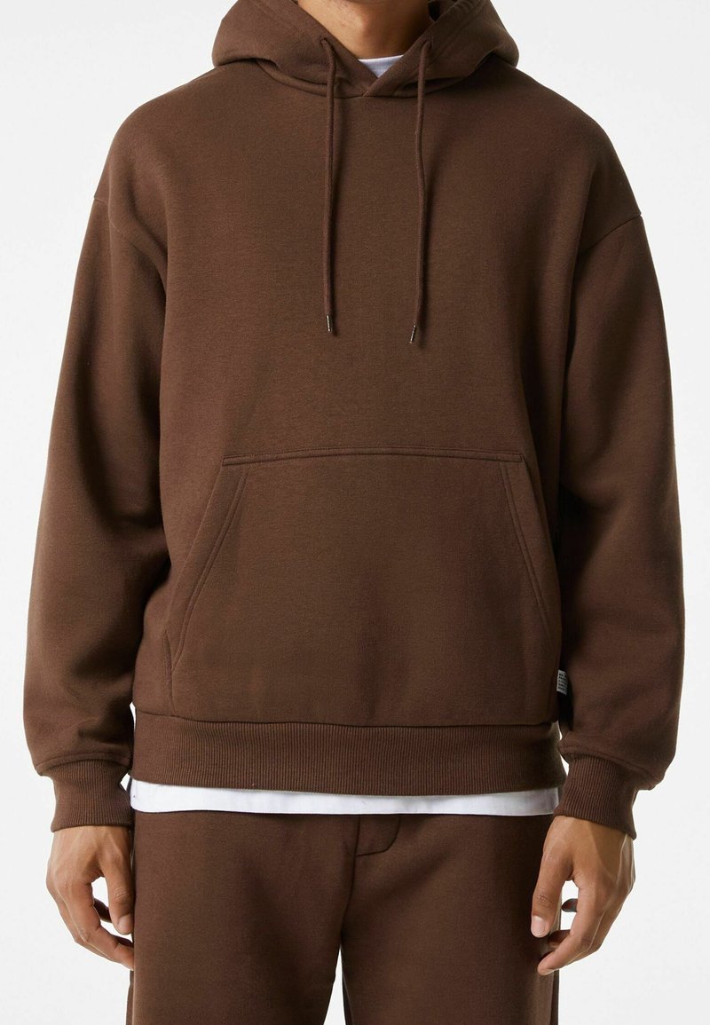 Homme portant un sweat à capuche marron uni avec une poche avant et un pantalon marron assorti, chemise blanche visible en dessous.