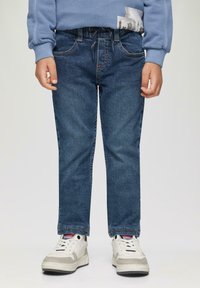 Donkerblauwe denim jeans met rechte pijpen, voorzien van voorzakken en subtiele stiksels. Gecombineerd met witte sneakers en een blauwe sweatshirt.