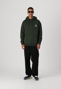 Sudadera verde oliva con bolsillo canguro, capucha y cordones, que presenta un logo triangular blanco. Combinada con pantalones negros holgados y zapatillas.