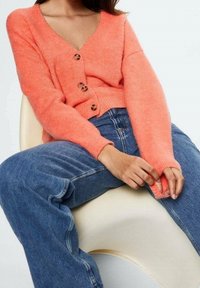 Femme assise sur une chaise blanche, portant un cardigan corail boutonné et un jean bleu, ajustant la manche du cardigan.
