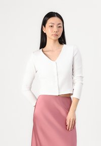 Cardigan blanc à côtes avec un col en V, des manches longues et une fermeture à boutons, associé à une jupe en satin rose. Texture lisse et design ajusté.