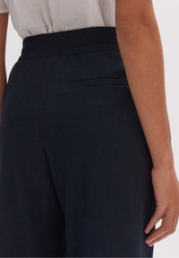 Tmavě modré kalhoty s texturovanou tkaninou, elastickým pasem a zadní kapsou. Střih je široký a volný.