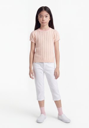 Fiatal lány világos rózsaszín rövid ujjú kötött felsőben, fehér rövidnadrágban, rózsaszín zokniban és fehér slip-on cipőben áll fehér háttér előtt.