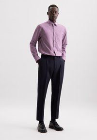 Mann steht mit den Händen in den Taschen, trägt ein pink gestreiftes Langarmhemd, dunkle Hosen, schwarze Socken und schwarze Anzugschuhe.