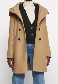 Camel uldblanded frakke med bred krave, dobbeltknaplukning foran, sorte knapper og sidelommer, båret over en sort cardigan og jeans.