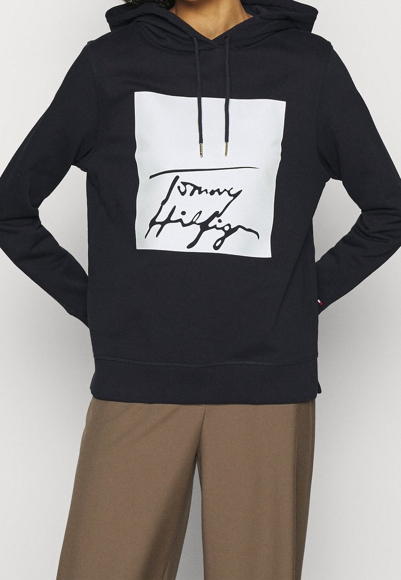 Personne portant un sweat à capuche noir avec un carré blanc et un texte manuscrit noir sur la poitrine, associé à un pantalon marron.