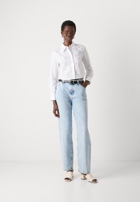 Blusa bianca con dettagli in pizzo, jeans azzurri dritti e sandali beige con tacco. Una cintura nera mette in risalto la vita.
