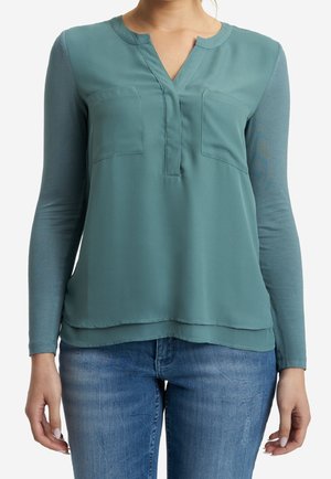 Femme portant un chemisier à manches longues couleur sarcelle avec deux poches poitrine et un jean bleu, debout les bras détendus le long du corps.