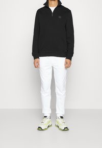 Hombre vistiendo un sudadera negra con cremallera hasta el cuarto, pantalones blancos y zapatillas blancas con detalles verdes, de pie contra un fondo claro y sencillo.