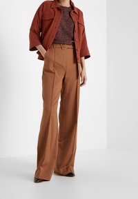 Veste couleur rouille avec de larges revers et deux poches poitrine sur un pull texturé foncé, associée à un pantalon marron taille haute à jambes droites.