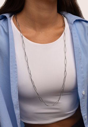 Lange zilveren ketting met langwerpige ovale schakels, gedragen over een witte top, aangevuld met een lichtblauwe overhemdbloes.