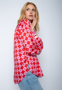 Ein rot und pink gemustertes Hemd mit geometrischen Designs, langen Ärmeln, seitlichen Schlitzen und gerippten Manschetten. Leichter Stoff, lockere Passform.