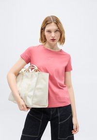 Νεαρή γυναίκα που φοράει ροζ μπλουζάκι και σκούρο παντελόνι κρατά μια μεγάλη κρεμ τσάντα ώμου μπροστά σε ένα απλό ανοιχτό γκρι φόντο.