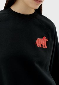 Zwarte sweatshirt met een rode gestreepte berenborduring op de borst. De stof lijkt zacht en heeft een geribbelde kraag en raglanmouwen.