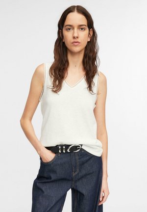 Kobieta z długimi brązowymi włosami, ubrana w biały bezrękawnik z dekoltem w serek, wpuszczony w ciemnoniebieskie dżinsy z czarnym paskiem, stojąca z jedną ręką w kieszeni.