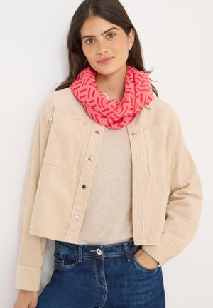 Beige Cordjacke mit silbernen Knöpfen, über einem hellen Pullover getragen, kombiniert mit einem rosa gemusterten Infinity-Schal, vor einem weißen Hintergrund.