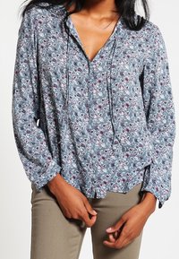 Blouse bleue à fleurs avec un col en V, des manches longues et une coupe décontractée. Présente un motif délicat avec des nuances de rose et de blanc.
