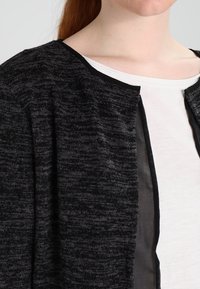 Svart och grå melerad cardigan med en strukturerad stickning, öppen front och genomskinliga accenter längs kanterna. Lätt och avslappnad passform.