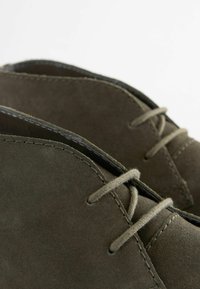 Zapatos de ante verde oliva con punta redondeada, costuras sutiles y cordones a juego, que muestran una textura suave y un diseño minimalista.
