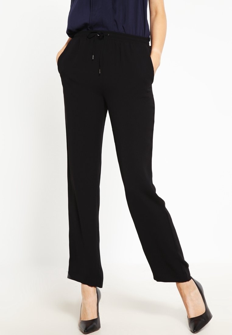Femme portant un pantalon noir à cordon de serrage et des talons noirs, debout les mains dans les poches sur un fond blanc.