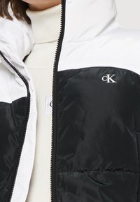 Žena oblečená v černo-bílé péřové bundě na zip přes krémový žebrovaný rolák se viditelnými logy Calvin Klein.