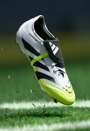 Bottine de football blanche avec des rayures noires et une semelle jaune éclatante. Présente un dessus texturé et une sangle sur les lacets pour un maintien sécurisé.