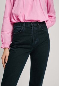 Jeans de mezclilla negra de cintura alta con un corte clásico, que cuentan con cinco bolsillos y un lavado oscuro. Combinados con una blusa rosa claro.