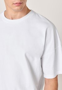 Bela bombažna majica z okrolim izrezom in kratkimi rokavi. Tkanina je videti gladka, z udobnim krojem in minimalnimi šivi.