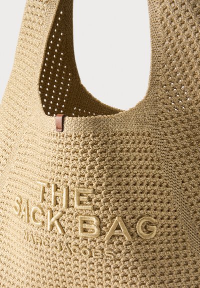 Sac fourre-tout beige en maille avec une large bandoulière et un motif texturé, brodé de "THE SACK BAG MARC JACOBS" sur le devant.