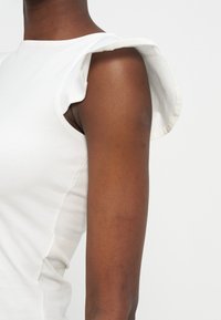 Κοντινό πλάνο στο πάνω μέρος του βραχίονα ενός ατόμου που φοράει λευκό αμάνικο τοπ με λεπτομέρεια βολάν στον ώμο πάνω σε ανοιχτό φόντο.