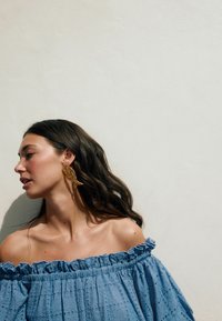Náušnice v tvare zlatých rýb s jemnými vzormi. Modelka má na sebe svetlomodrý top s odhalenými ramenami, s volánikmi a detaily z vyšívanej látky.