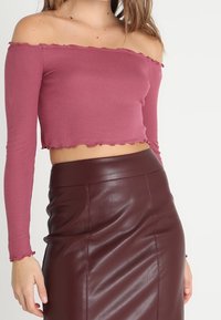 Off-the-shoulder, long-sleeve top in mauve ribbed fabric with wavy hemline, paired with a high-waisted burgundy faux leather skirt.