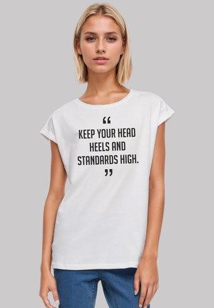 Weißes Baumwoll-T-Shirt mit umgeschlagenen Ärmeln, mit fettem schwarzen Text, der lautet: "HALTE DEINEN KOPF, DEINE FÜßE UND DEINE STANDARDS HOCH." Lässige Passform.