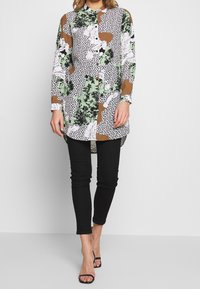 Langærmet skjorte med et blomster- og polkaprikkemønster i grøn, sort, hvid og brun, parret med sorte slim-fit bukser og sandaler med stropper.