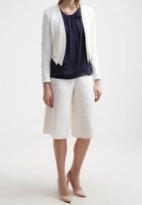 Weißer, kurzer Blazer über einer marineblauen Bluse, kombiniert mit weißen Culottes und nudefarbenen High Heels. Glatte Texturen und maßgeschneiderte Designelemente sind deutlich sichtbar.