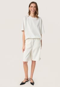 Hvid bomulds t-shirt med korte ærmer parret med hvide denimshorts. Modellen har sorte spidse flade sko og minimale sølv-øreringe i hoop-stil på.