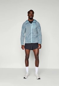 Lichtblauwe, lichte jas met rits en een voorzak, gedragen over een zwart shirt en shorts. Heeft een capuchon en elastische manchetten.