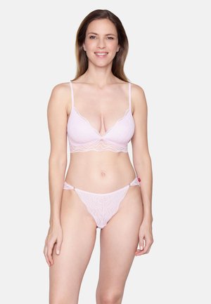Femme debout et souriante, portant un soutien-gorge en dentelle rose clair avec des bretelles fines et une culotte assortie en dentelle, sur un fond clair uni.