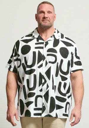 Mann mit kurzen Haaren und Bart trägt ein weißes kurzärmliges Hemd mit kräftigen schwarzen abstrakten Formen und beigen Hosen auf schlichtem Hintergrund.