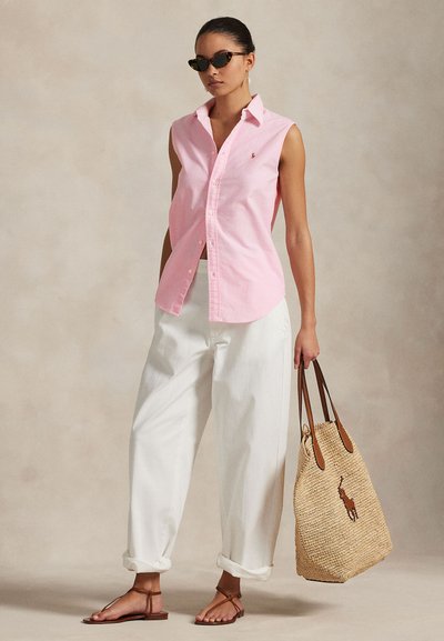 Chemise sans manches rose clair avec boutons, pantalon blanc décontracté, sandales plates marron, et sac tressé beige avec accents foncés et détail logo.