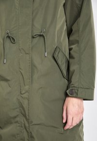 Veste vert olive avec cordons à l'avant, poche à rabat inclinée et poignets boutonnés sur la manche droite, portée par une personne dont la main droite est visible.
