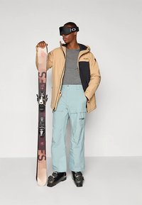 Άνδρας που φορά μπεζ μπουφάν, γκρίζο πουκάμισο, ανοιχτό μπλε παντελόνι για σκι, μαύρες μπότες για σκι και μαύρα γυαλιά σκι, κρατώντας ένα όρθιο σκι.