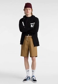 Black zip-up hoodie, black t-shirt, beige shorts, and black sneakers with blue accents. Model wears a maroon beanie and white socks with logos.