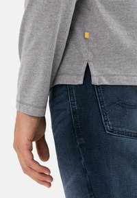 Grå langærmet skjorte med rundet kant, sidespalte og et lille gult mærke. Bærende sammen med mørke denim jeans, der har en afslappet pasform.