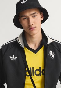 Sombrero tipo bucket negro con logo blanco, chaqueta negra con cremallera y detalles blancos con logo, camiseta amarilla con cuello en v y texto negro en negrita, con gráfico de caballo.