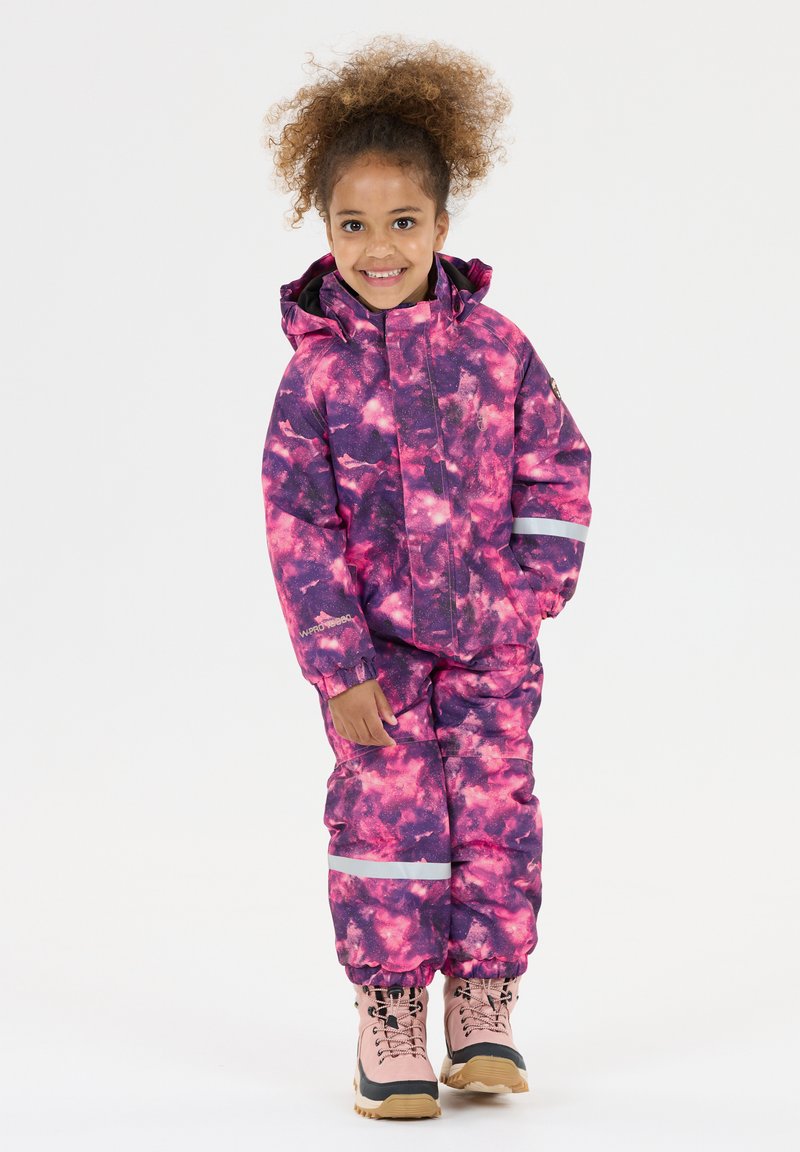 Smilende barn iført en vandtæt snowsuit i pink og lilla med reflekterende bånd og lyserøde vinterstøvler på en hvid baggrund.
